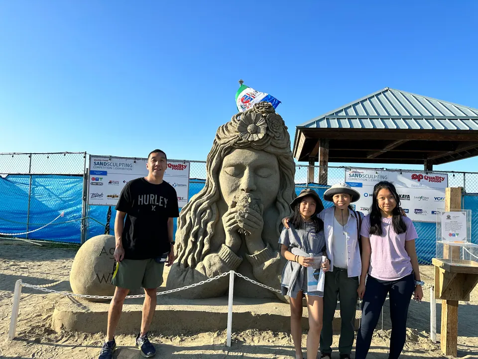 Four people stand in front of a tall sand sculpture of a woman's face with flowers in her hair, under a blue sky.
