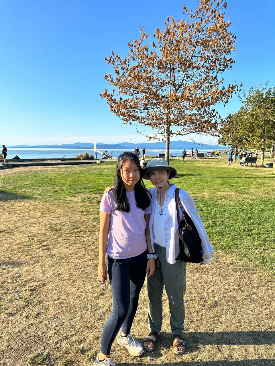 Two people stand on a sunny lawn by the sea, with a tree and a blue sky behind them.