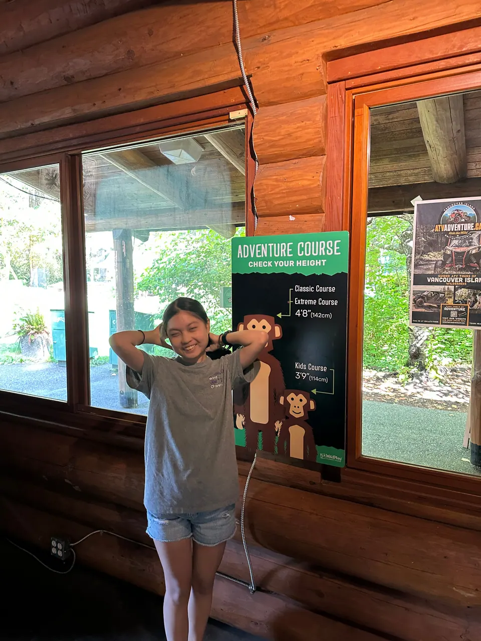 A girl poses with her hands on her head beside a green Adventure Course sign inside a log building.