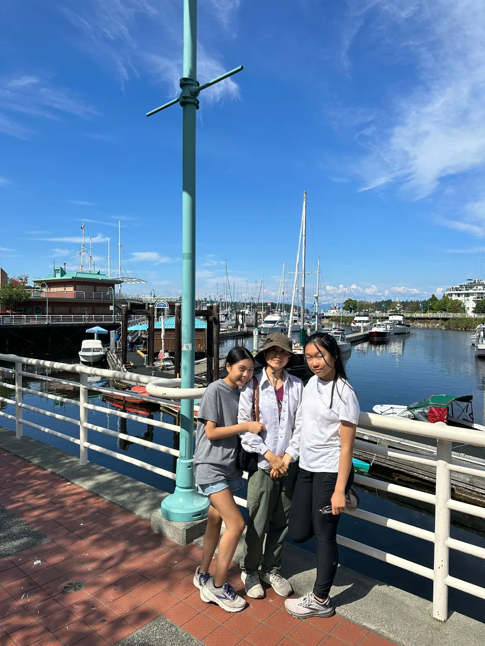 Three people stand at a railing on a waterfront walkway beside a marina under a blue sky.