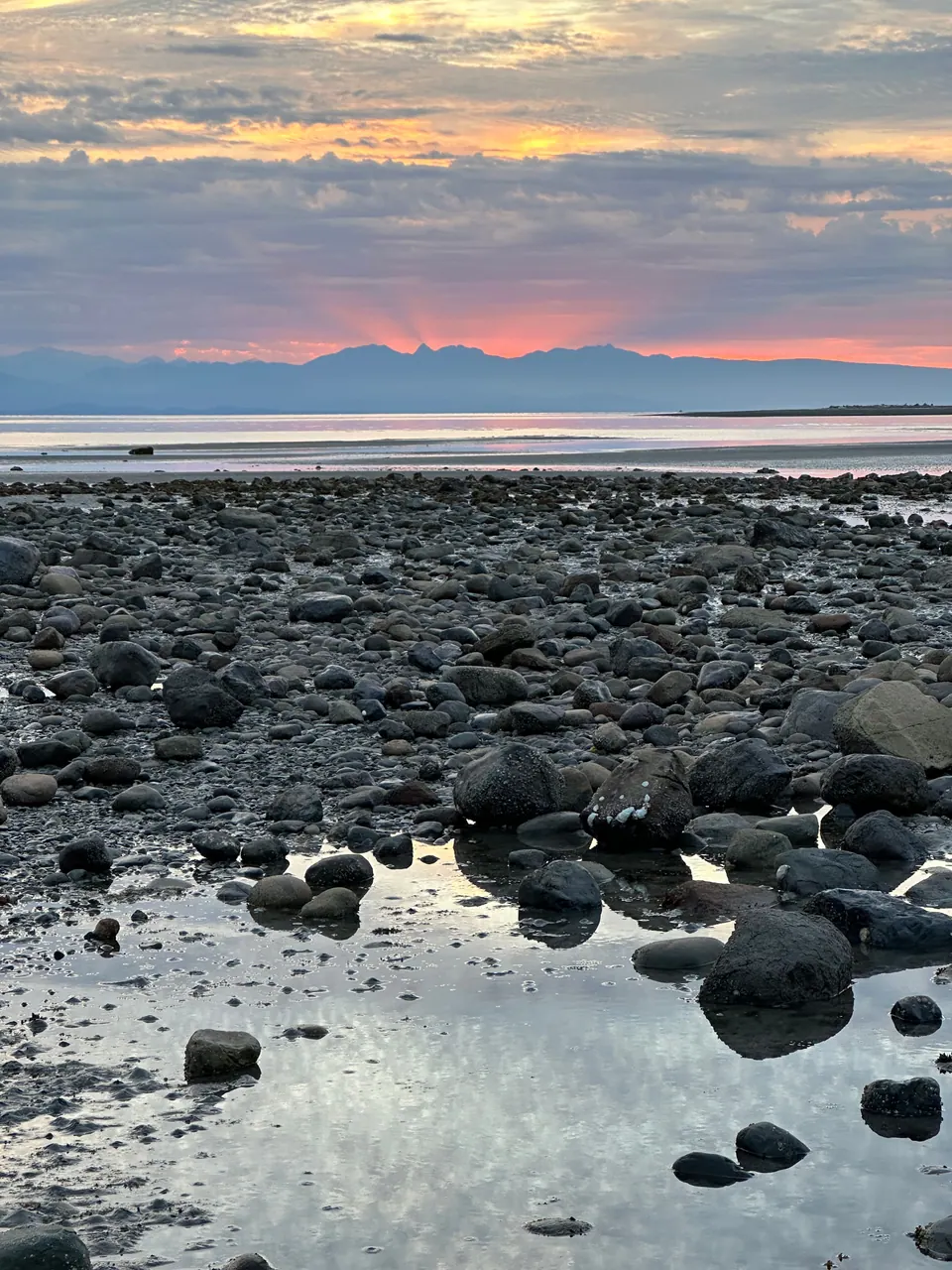 A pink and orange sky over distant mountains above a wide, stony tidal beach.