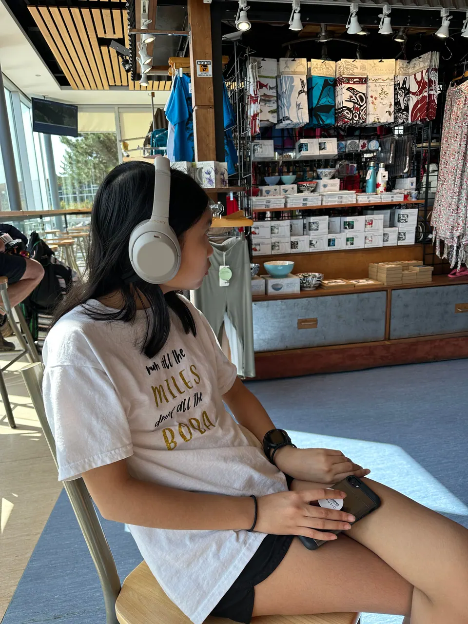 A girl in headphones sits in a chair in a bright building with a gift shop behind her.