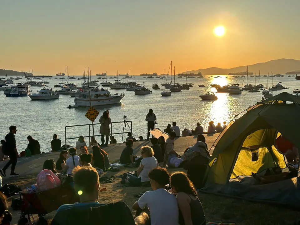 A crowded beach at sunset with tents and people, and many boats anchored in the bay under an orange sun.