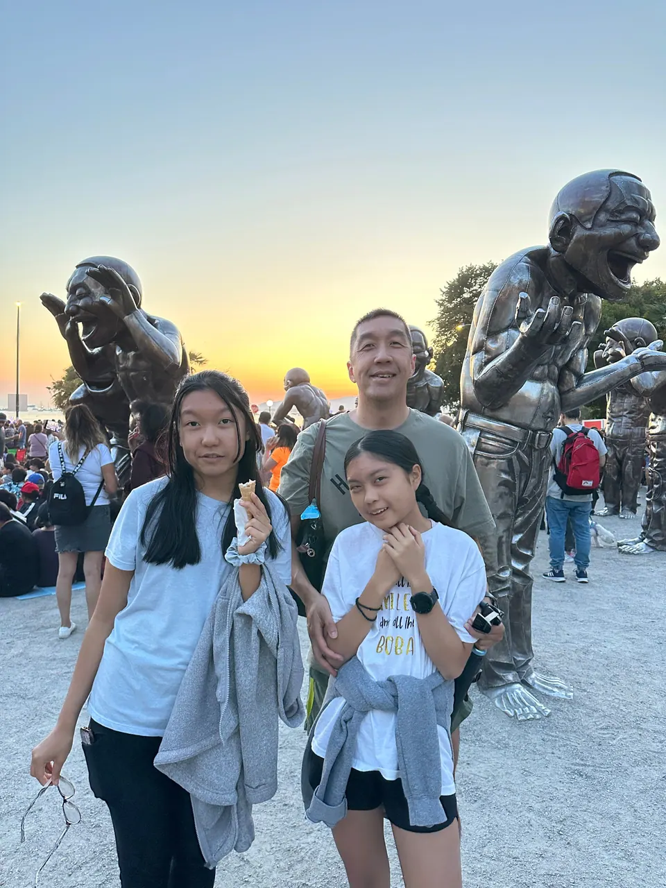 Two girls and a man stand among large bronze statues of laughing men, with an orange sky behind.