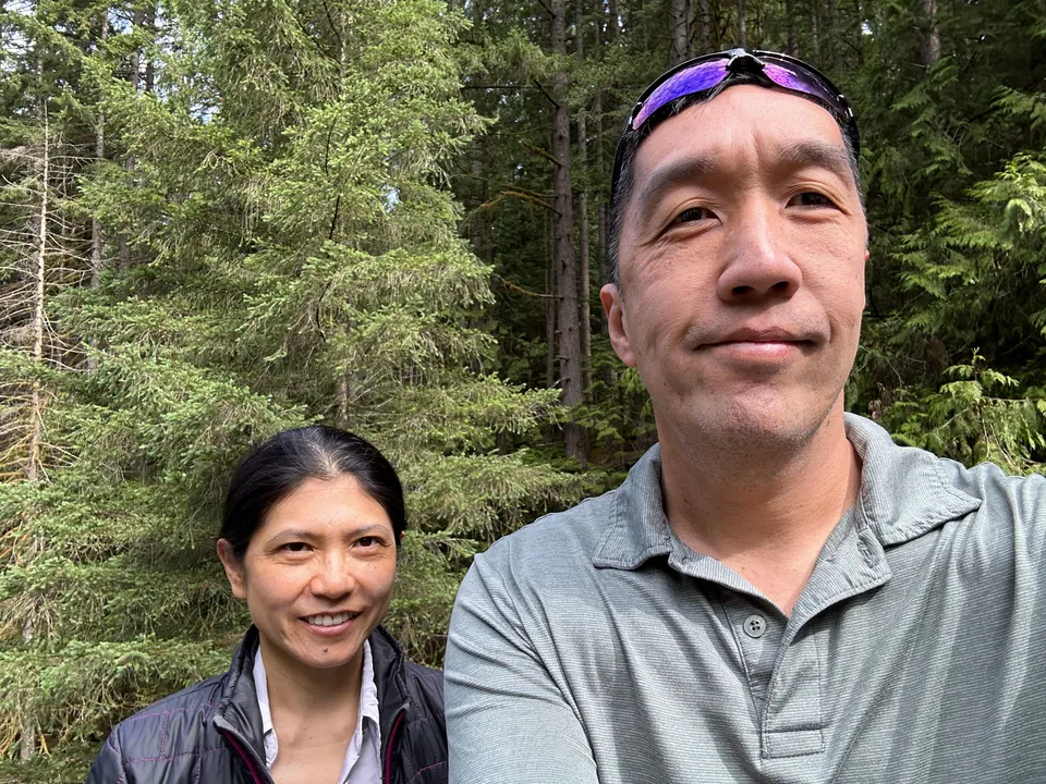 A man and a woman take a selfie in front of tall evergreen trees.