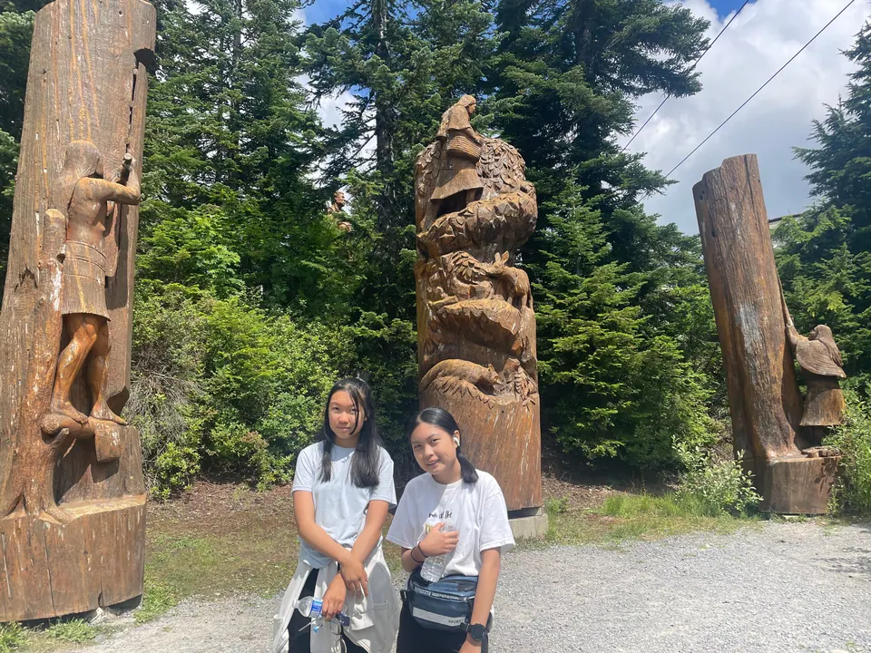 Two girls stand in front of tall wooden carvings of figures and animals in a forest clearing.
