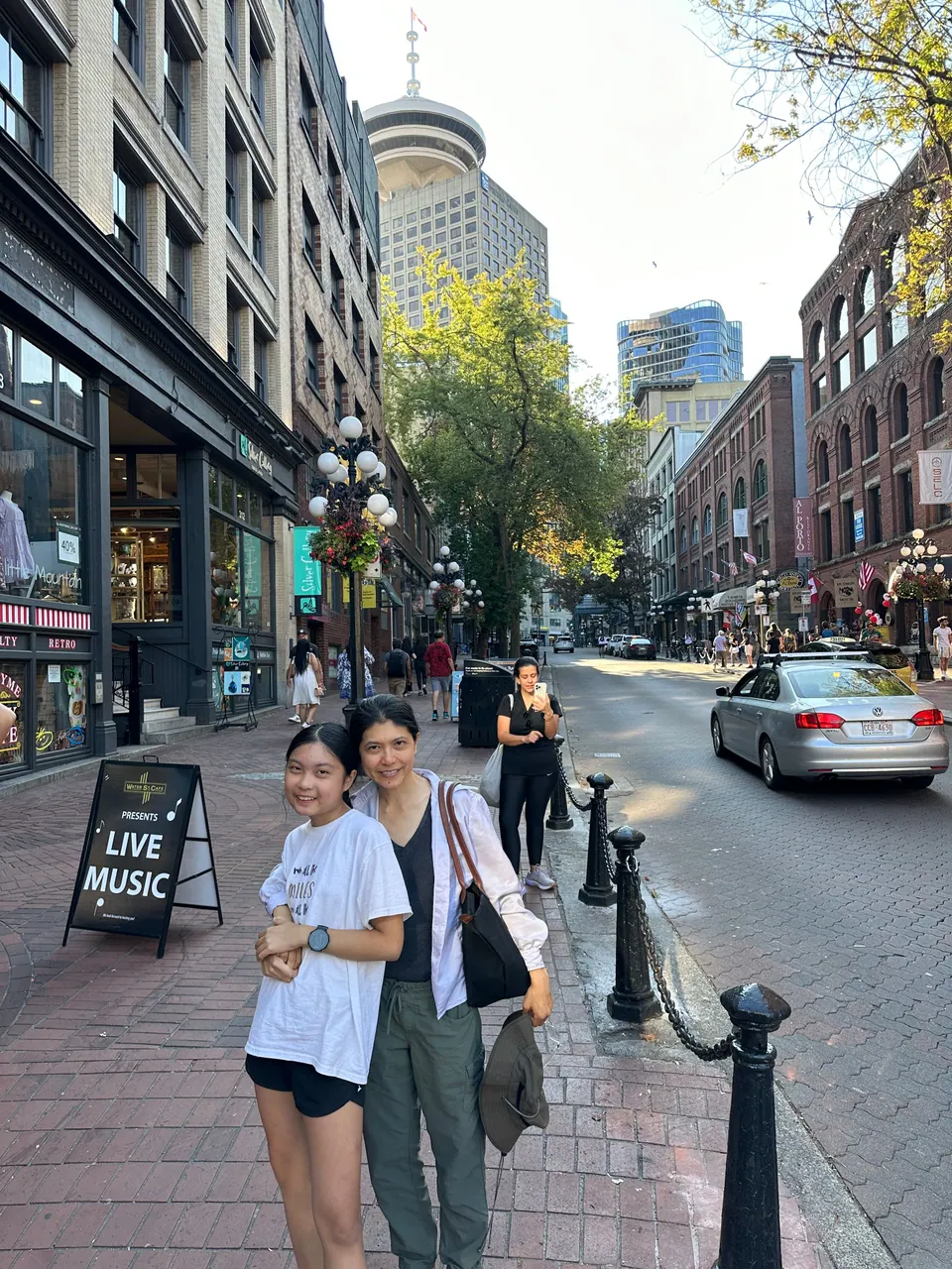 Two people pose on the brick sidewalk of an old street lined with brick buildings; a tall tower with a round top is behind them.