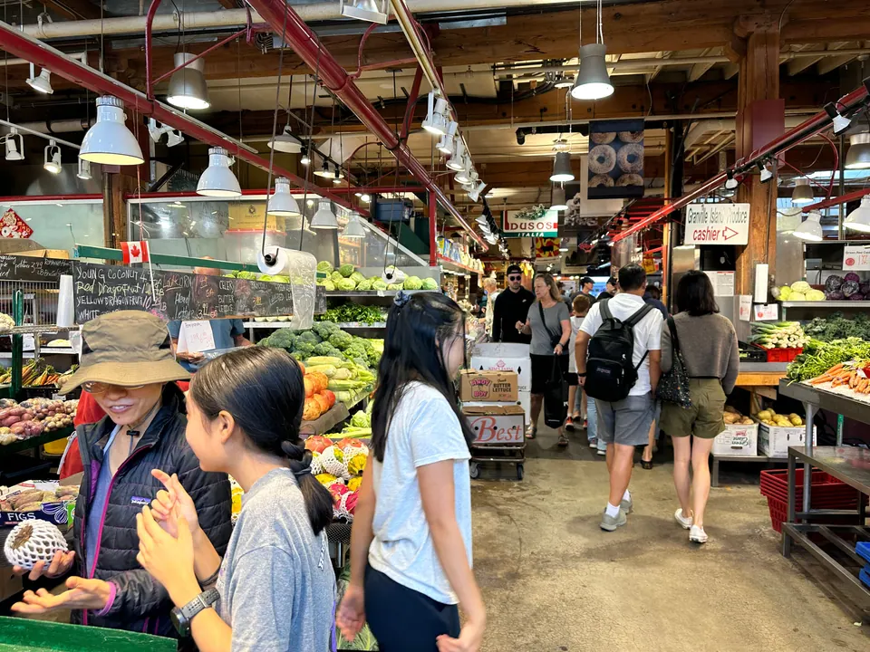Shoppers walk between produce stalls under industrial beams in a busy indoor market.