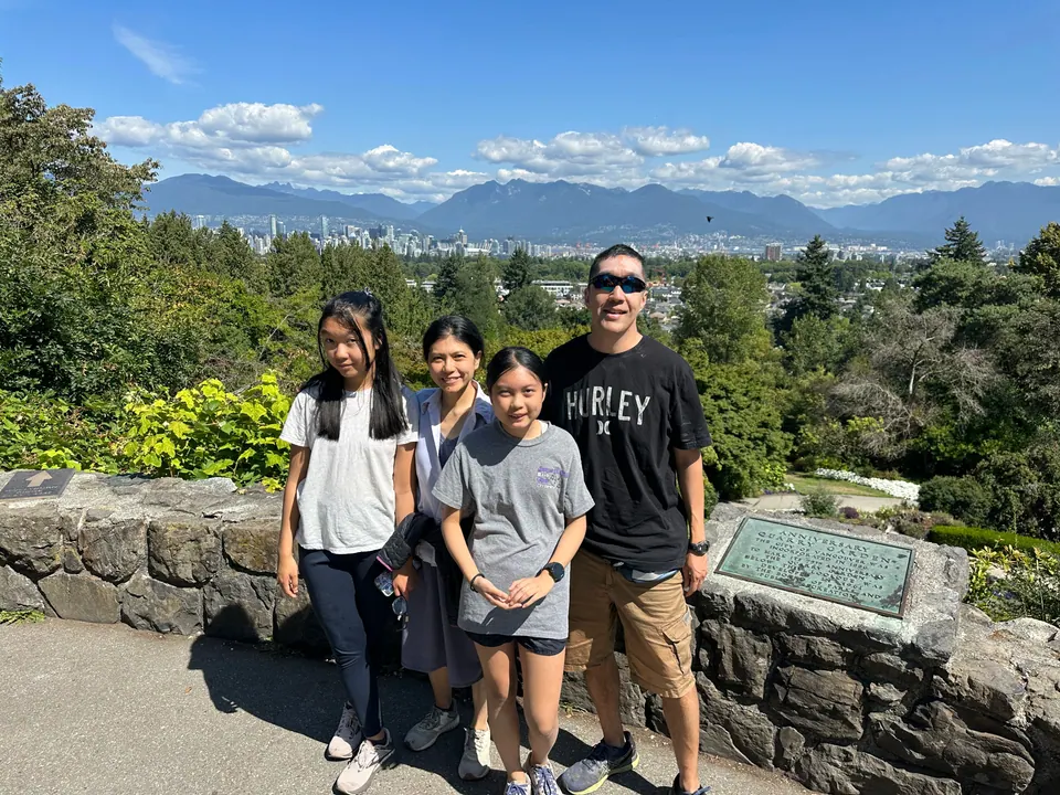 Four people stand at a stone wall on a hilltop, with a city, forest and mountains behind them under a clear sky.