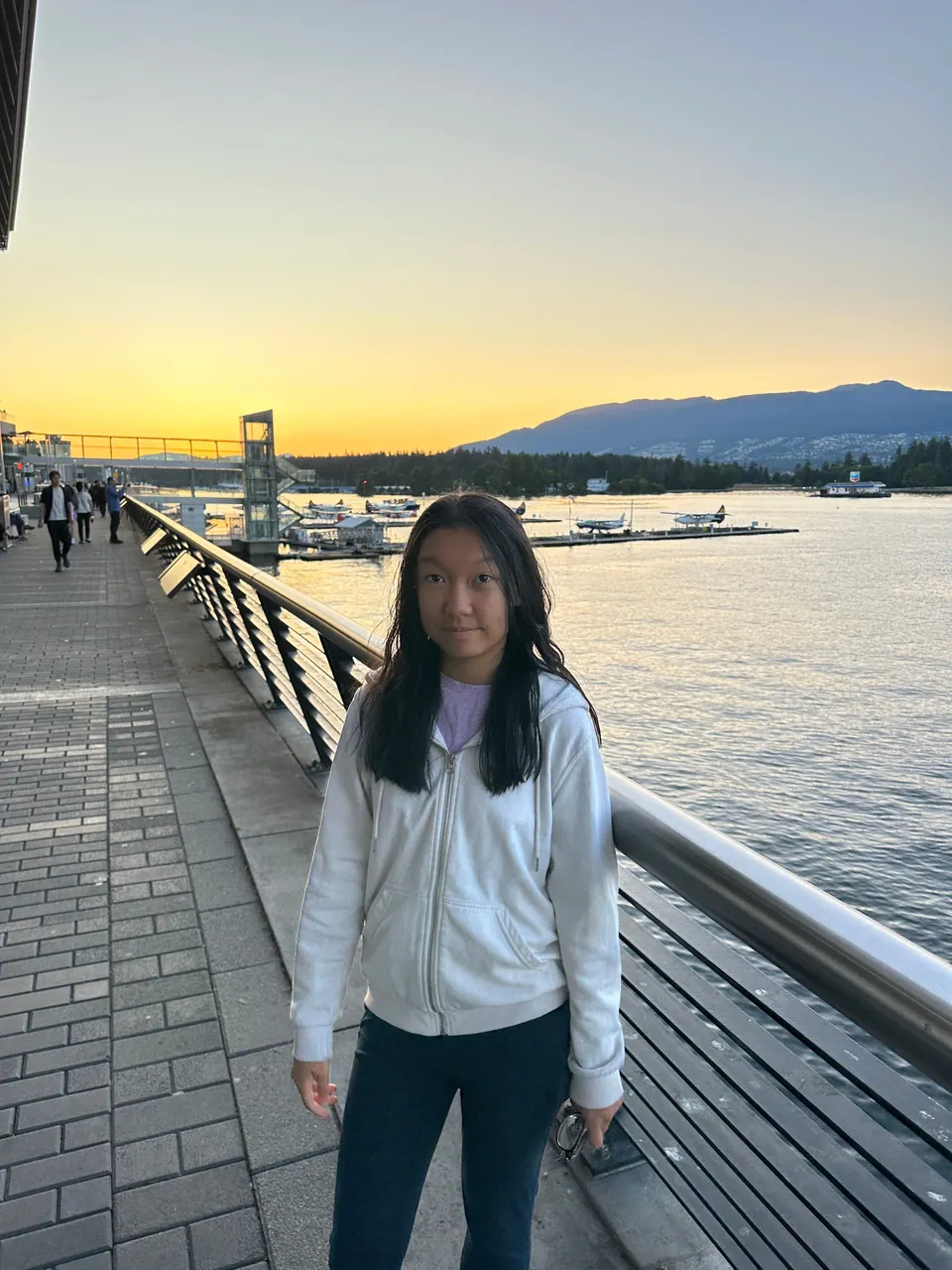 A girl in a white jacket stands on a waterfront boardwalk at sunset, with mountains across the water.