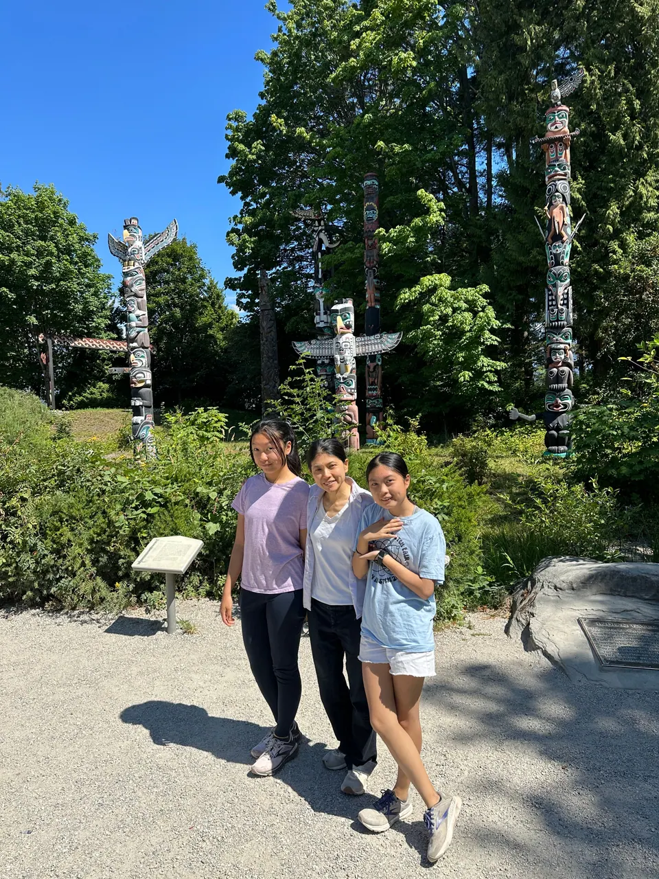 Three people pose on a gravel path in front of tall carved and painted totem poles and trees.