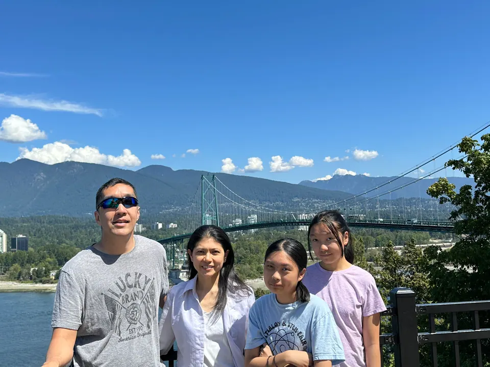 Four people smile at a lookout; behind them a green suspension bridge crosses to forested mountains under a blue sky.