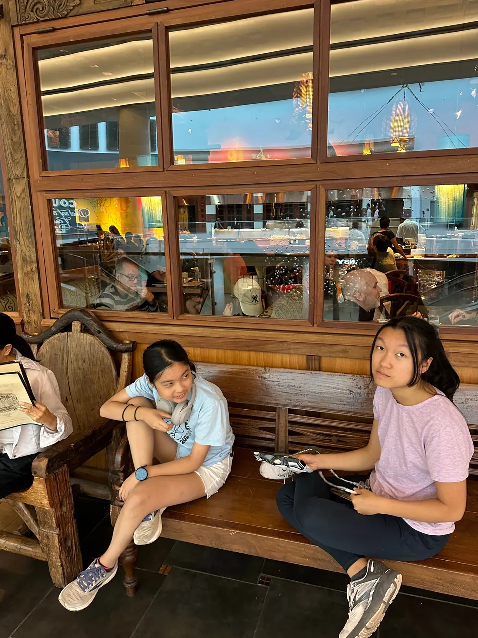 Two girls sit on a wooden bench in front of a glass display case.
