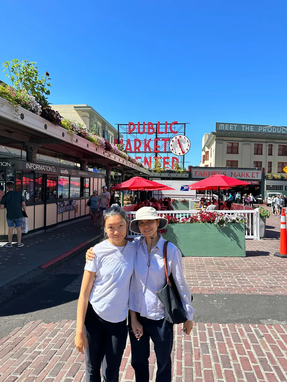 Two people pose on a brick street in front of the red Public Market Center sign and awnings.