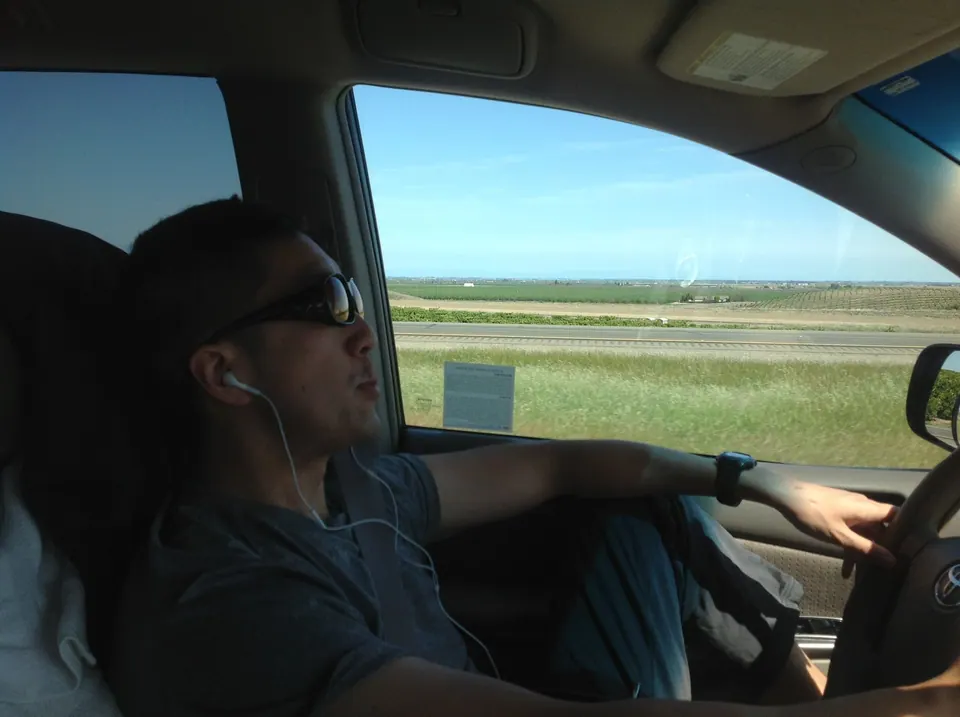 Seen from the passenger seat, a man in sunglasses and earbuds drives along a sunny freeway past flat green fields.