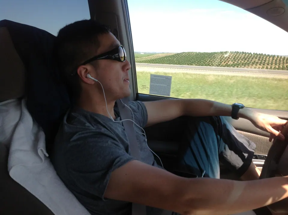 Seen from the passenger seat, a man in sunglasses and earbuds drives with one arm on the wheel; rows of orchard trees and a highway sign pass beyond the window.