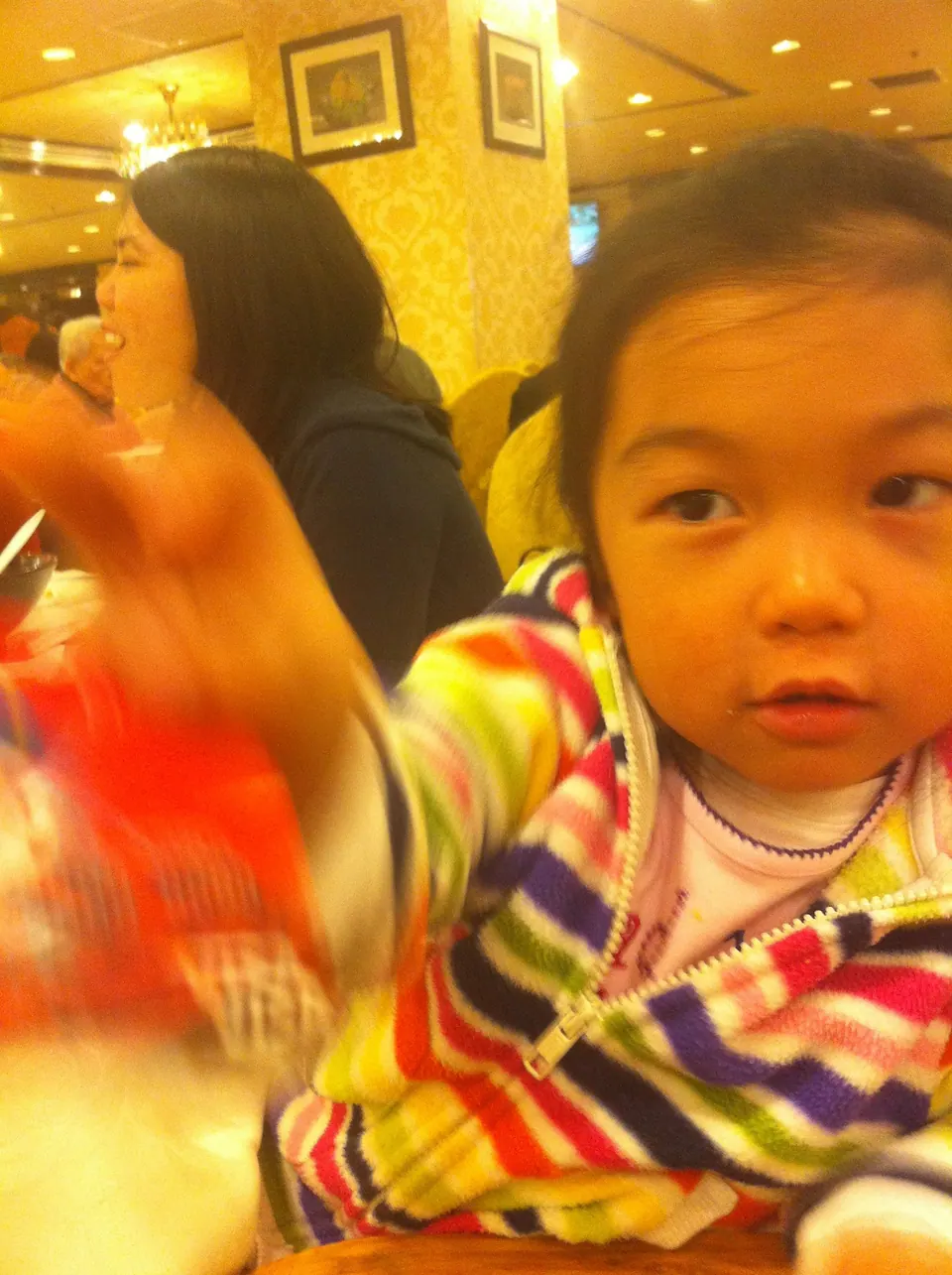 A toddler in a rainbow-striped hooded jacket looks at the camera across a restaurant table, one hand a blur; a woman sits eating behind her.