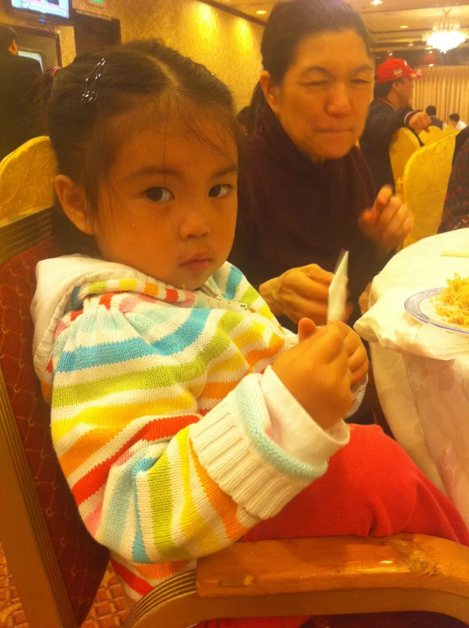 A little girl in a rainbow-striped hooded jacket sits in a red and gold banquet chair holding a napkin, with a plate of noodles on the round table and an older woman eating behind her.