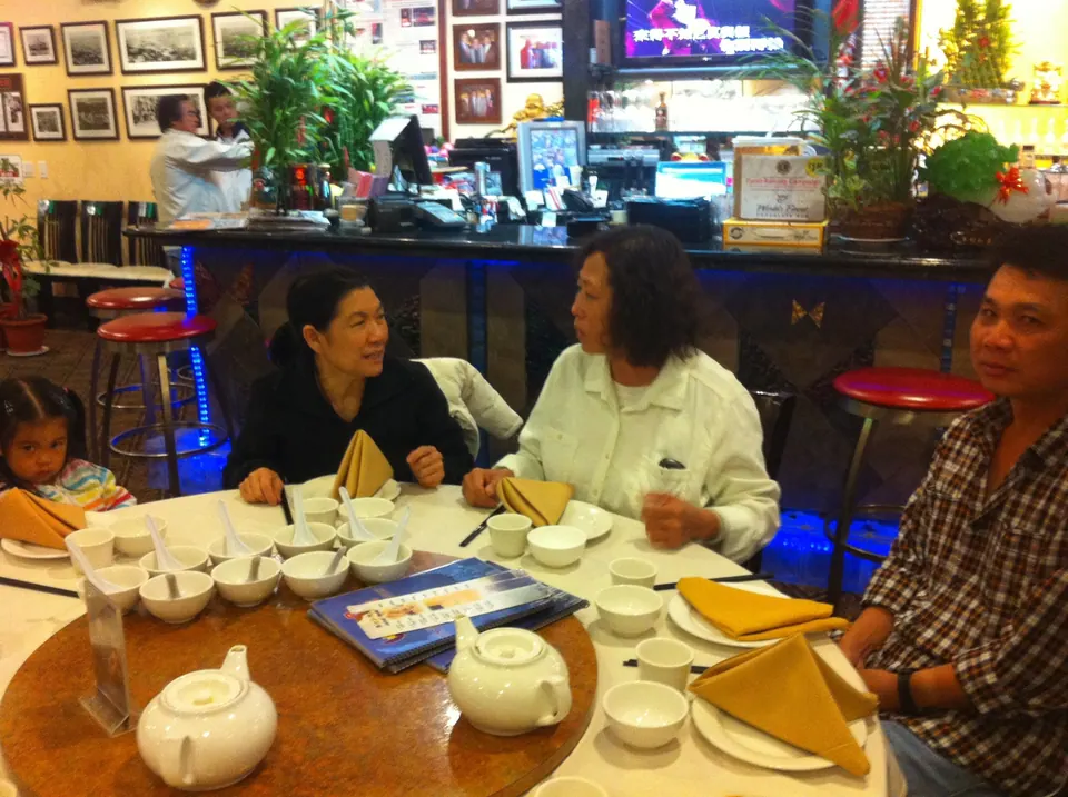 Around a round table with teapots and menus, an older woman in a white jacket talks with a woman in black and a man in a checked shirt; a small girl in a striped jacket peeks in at the left edge, with a lit bar and framed photos behind.