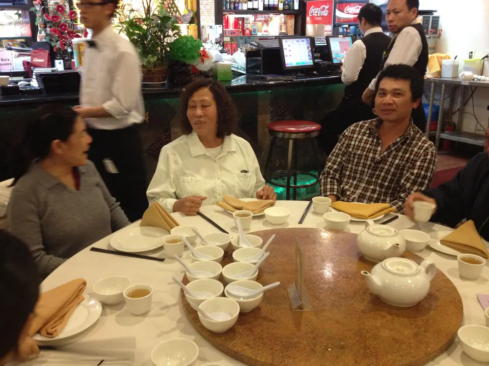A wide view across the round table, with rows of teacups and bowls and a lazy Susan, while a man in a checked shirt and an older woman in white talk and a waiter in a vest passes behind.