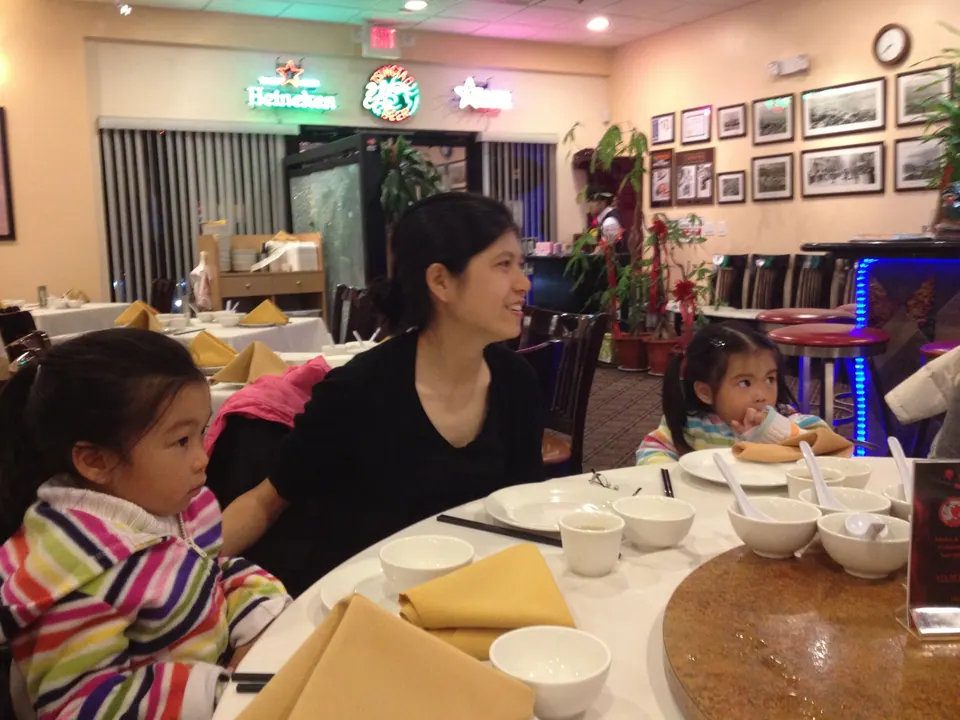 A woman in black smiles at the table beside two small girls in rainbow-striped jackets, with beer signs, framed black-and-white photos and empty tables behind them.