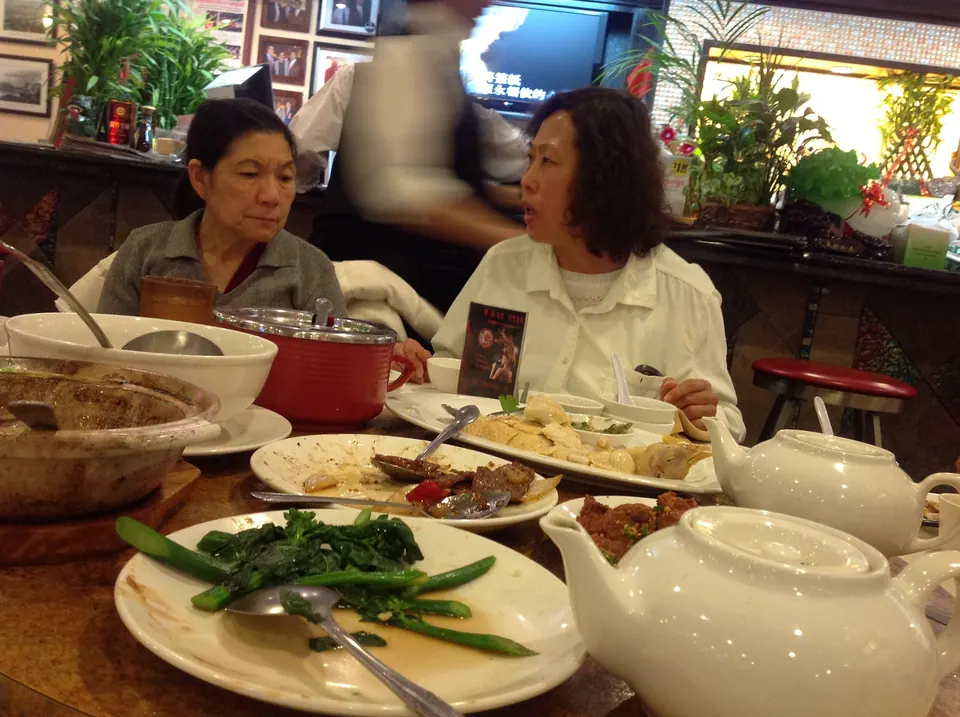 A red pot, a clay pot, a plate of chicken, gai lan and teapots crowd the lazy Susan while two women talk across the table.