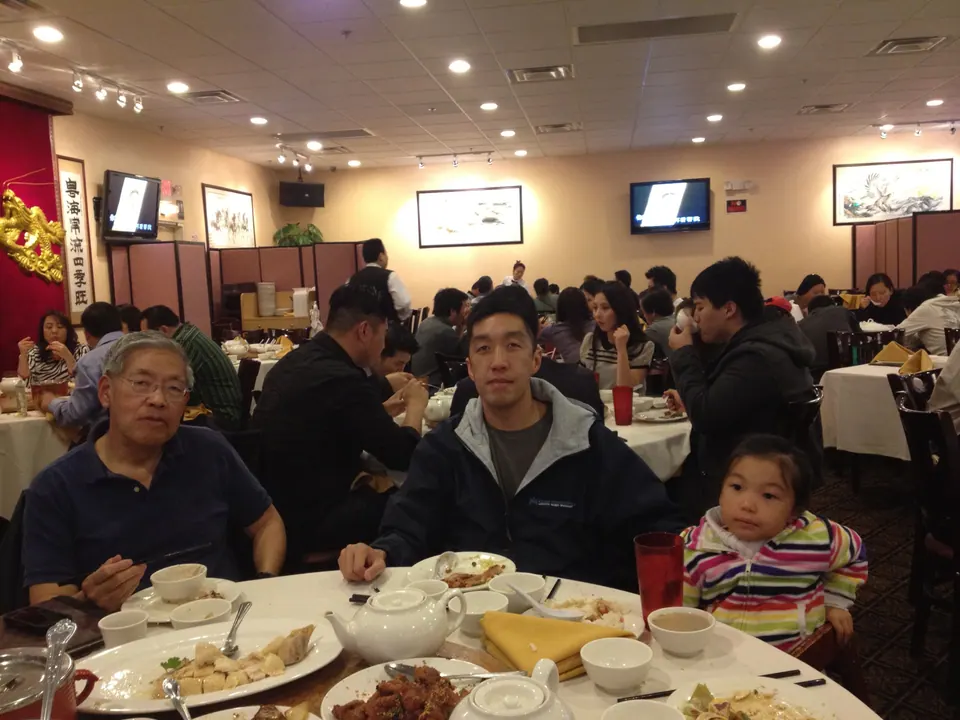 An older man in glasses, a young man in a dark jacket and a small girl in a rainbow-striped jacket sit at a round table of dishes in a busy restaurant with TV screens on the walls.