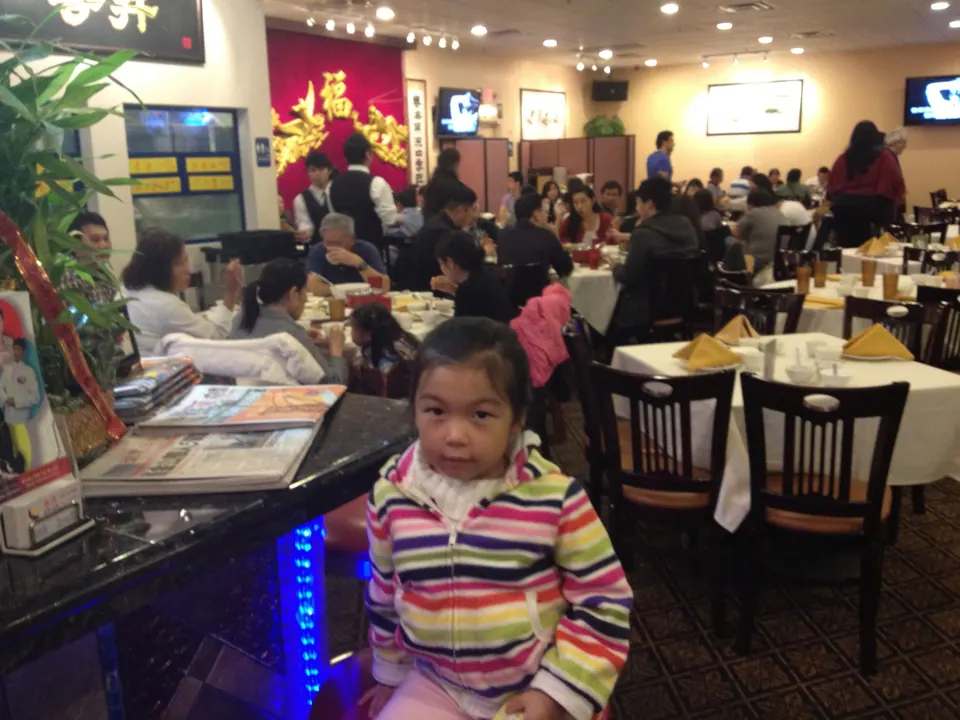 A small girl in a rainbow-striped jacket sits at the bar rail looking at the camera, with the busy dining room, a red banner and a stack of newspapers behind her.