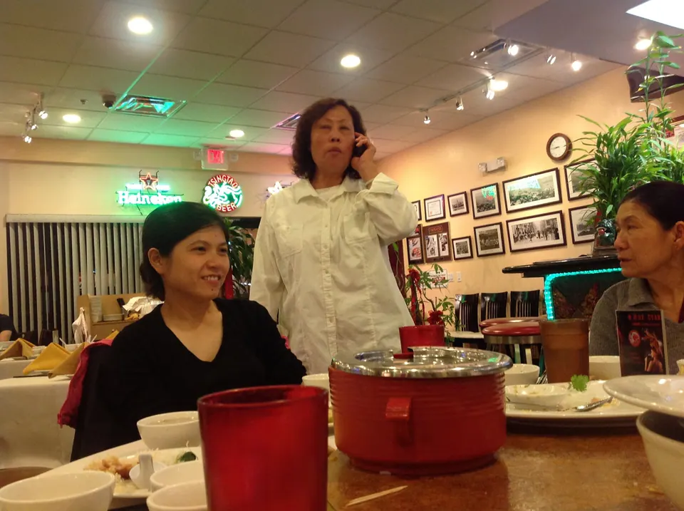 An older woman in a white jacket stands talking on a phone behind a smiling woman in black, with a red pot on the table and a Heineken sign glowing on the wall.