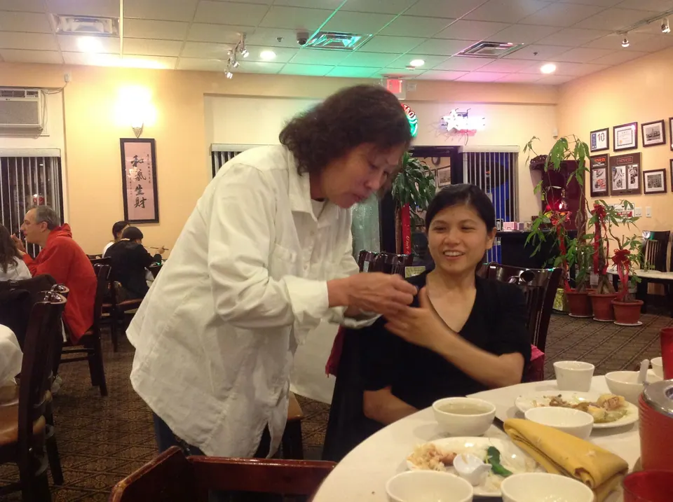 An older woman in white leans over to look at a phone held by a smiling woman in black, with the emptying table and quiet restaurant behind them.