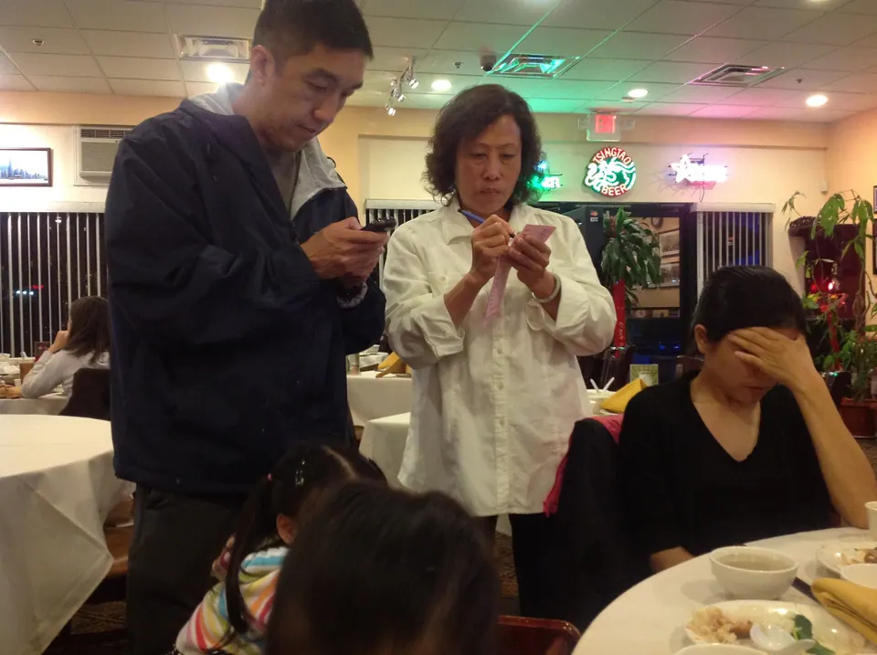 A man in a dark jacket and an older woman in white stand over their phones while a woman in black rests her forehead on her hand; the top of a child's head shows at the bottom of the frame.