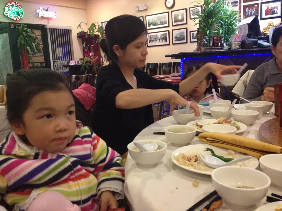 A girl in a striped jacket looks off to the side beside a woman in black reaching for a bowl; the table is covered in used plates and empty soup bowls.