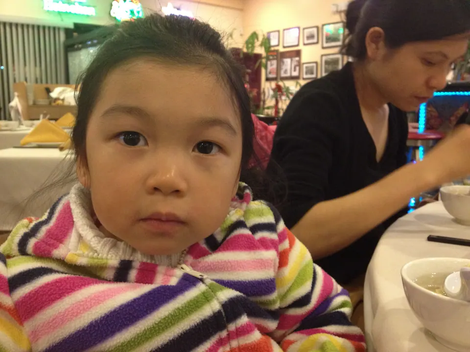 A close portrait of a little girl in a rainbow-striped fleece looking straight into the camera, with a woman in black eating beside her.