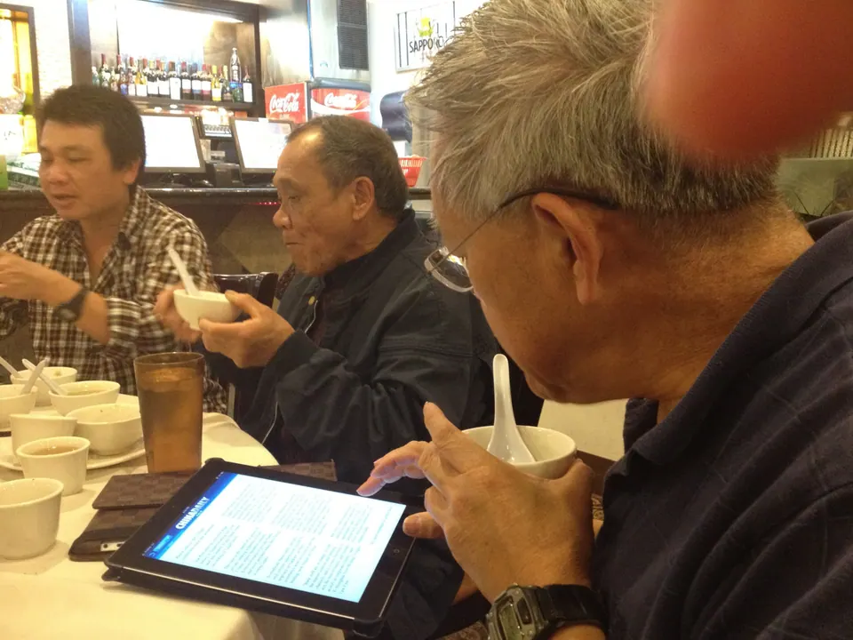 Three men at the table over bowls of soup: a man in a checked shirt talking, an older man in a dark jacket lifting a bowl, and a grey-haired man in glasses with an iPad in front of him.