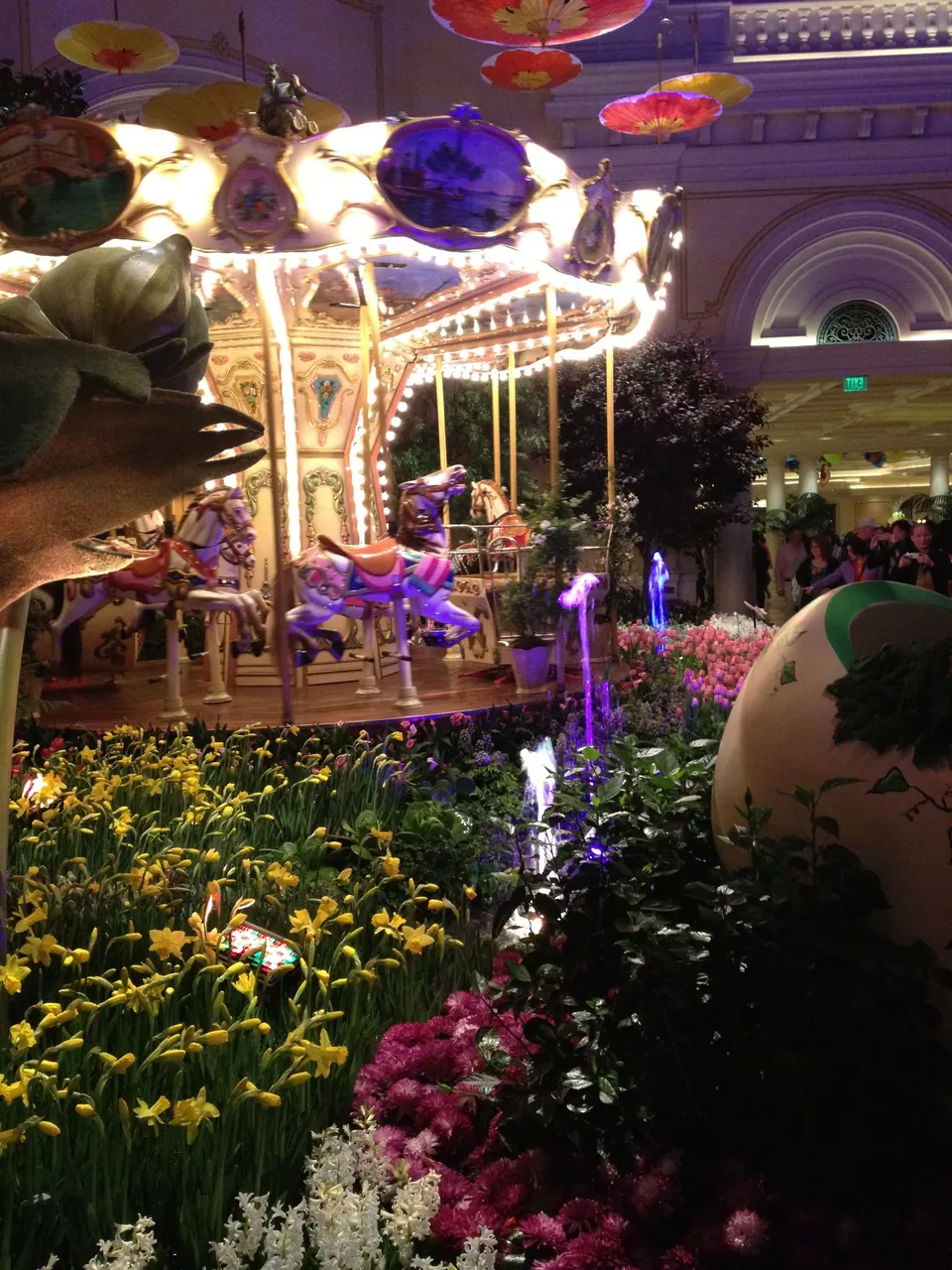 A lit carousel with painted horses glows behind beds of yellow daffodils and pink flowers, with a giant egg at the right.