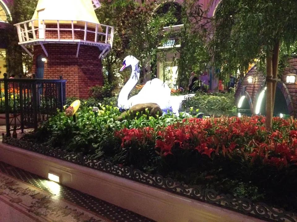 A white swan sculpture sits in front of the brick windmill tower and arched footbridge, above a bed of orange lilies.