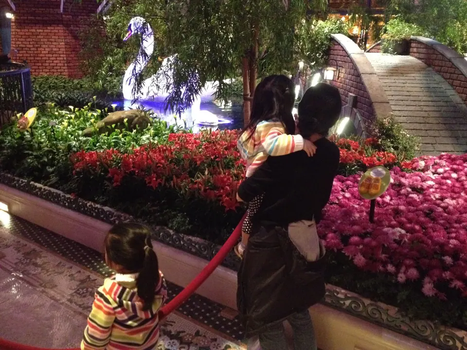 A woman in black carries a girl in a striped jacket and looks at the swan and brick footbridge, while a second girl in a ponytail stands at the rope.