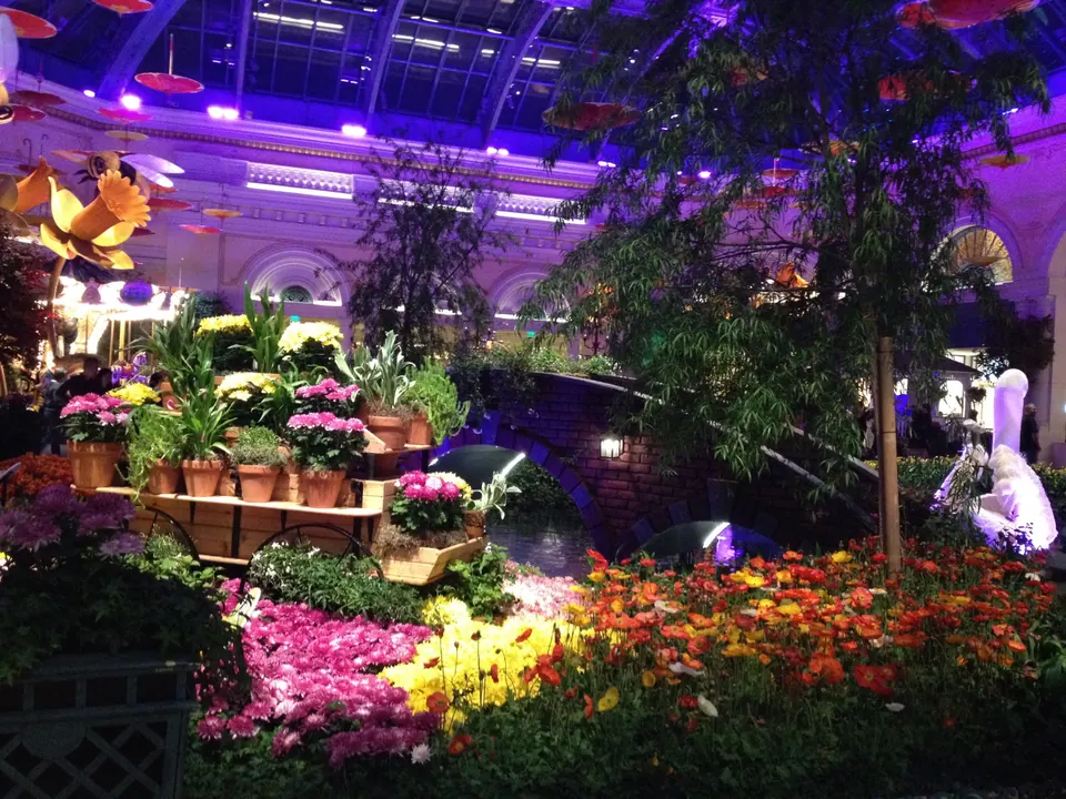 A garden cart of potted spring flowers stands beside the pond and brick bridge, with giant flowers and bees overhead and a swan glowing at the right.