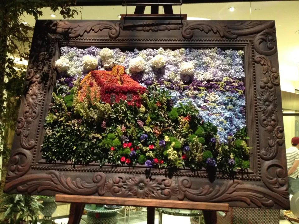 A big carved wooden frame on an easel holds a picture made entirely of flowers: a red house, hydrangeas and green hills.