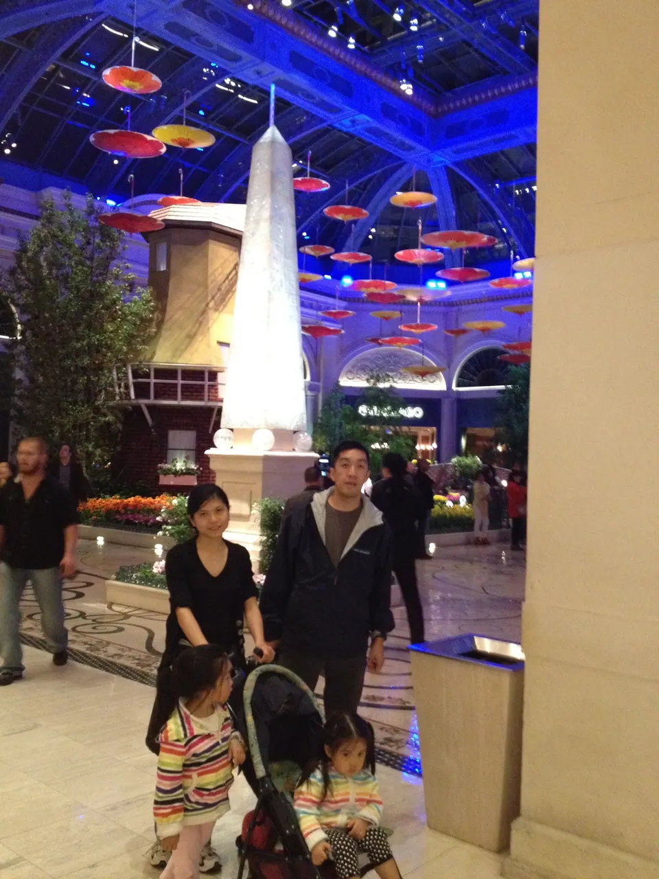 A woman and a man stand behind a stroller with two small girls in striped jackets, in a marble hall lit blue and purple, with parasols hanging from the ceiling.