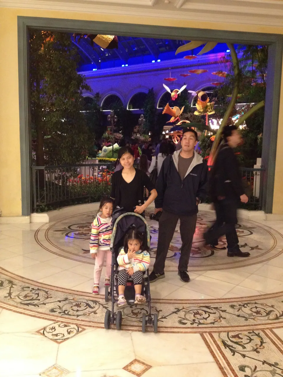 A man in a dark jacket and a woman in black stand behind a stroller with two girls in striped jackets on a marble floor inlaid with a medallion, the Conservatory glowing behind them.