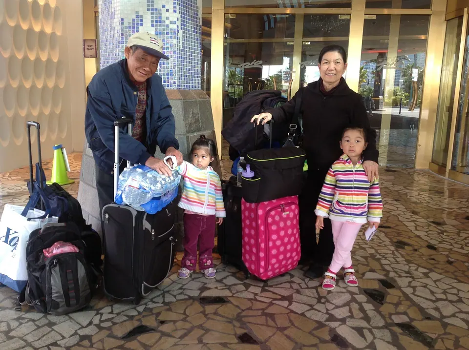 An older man in a cap and an older woman stand with two small girls in striped jackets beside a pile of suitcases, a pink rolling bag and a case of bottled water on a mosaic floor.