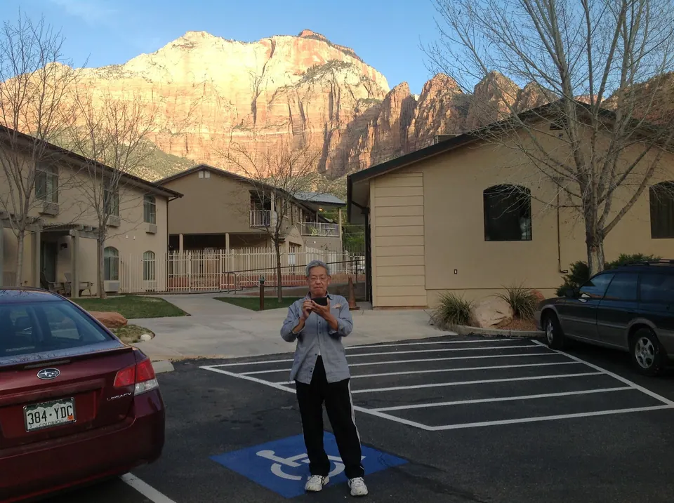 An older man in a grey jacket takes a photo in a motel parking lot; beyond the beige buildings a sunlit red and cream sandstone cliff rises into a clear sky.
