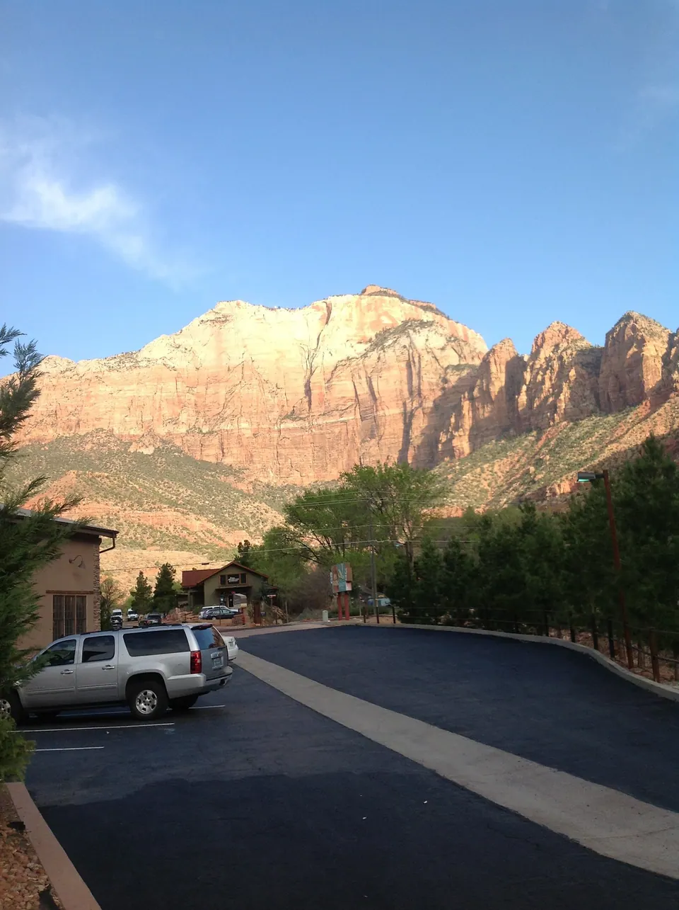 Sunlit cream and orange cliffs and buttes rise above the trees and motel roofs, seen from the parking lot in the early morning.