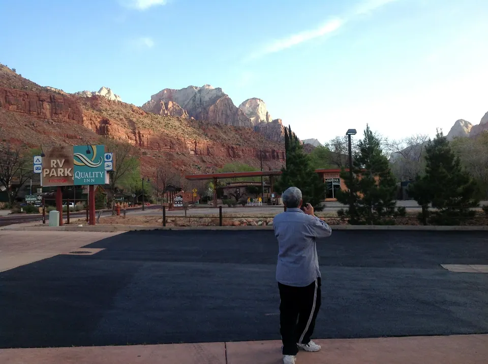 A grey-haired man in a grey jacket and track pants stands in the parking lot photographing the red cliffs, with the RV Park and Quality Inn sign behind him.