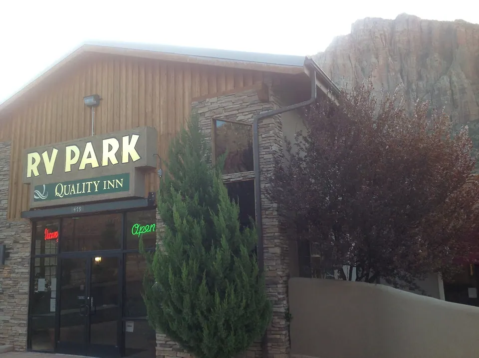 The wood and stone front of the motel with a lit "RV Park Quality Inn" sign, an open door and a red rock cliff above.