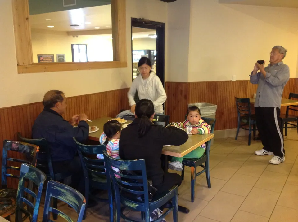 In a small breakfast room with wood paneling, an older man, a woman in black and two small girls sit at a table while a woman in a white jacket stands by and a grey-haired man photographs them.