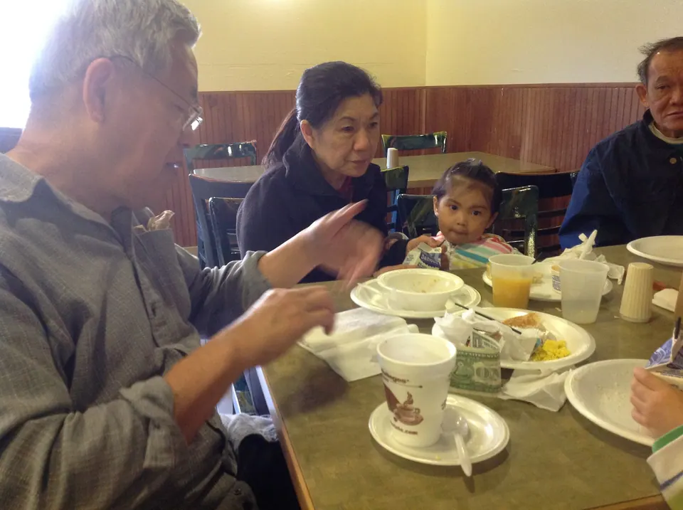 Around a table of foam cups and paper plates, a grey-haired man gestures while a woman and a little girl eat and an older man looks on.