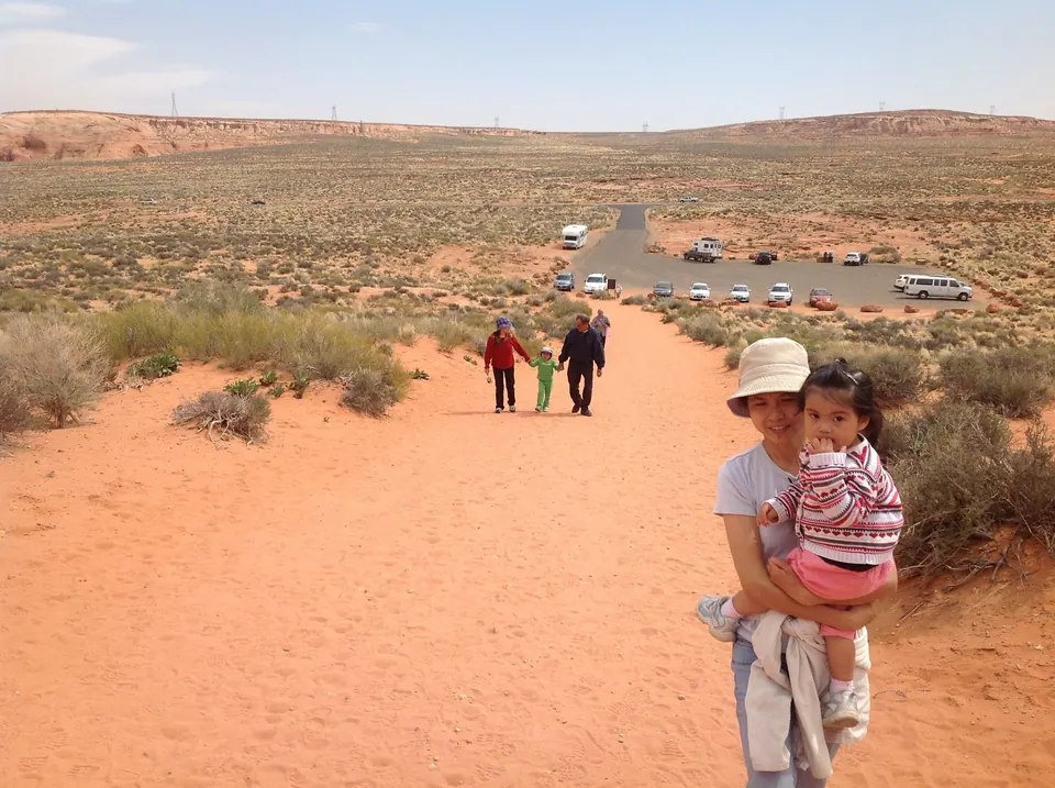 A woman in a white sun hat carries a little girl up a wide trail of orange sand through desert scrub; other walkers and a parking lot are behind them.