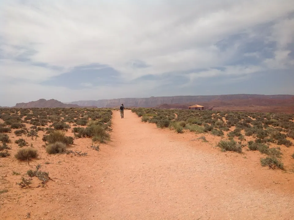 A wide straight sand trail through low sagebrush with a lone walker ahead, a small shade shelter and red cliffs in the haze.